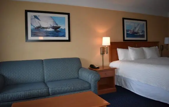 A teal upholstered sofa in the foreground of a room with a king bed behind it, two framed sailboat prints on the wall, and a nightstand with a phone