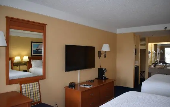 A hotel room with a wall-mounted flat-screen TV, a wood dresser holding a coffee maker, and a mirror reflecting a bed with white linens against a warm tan wall