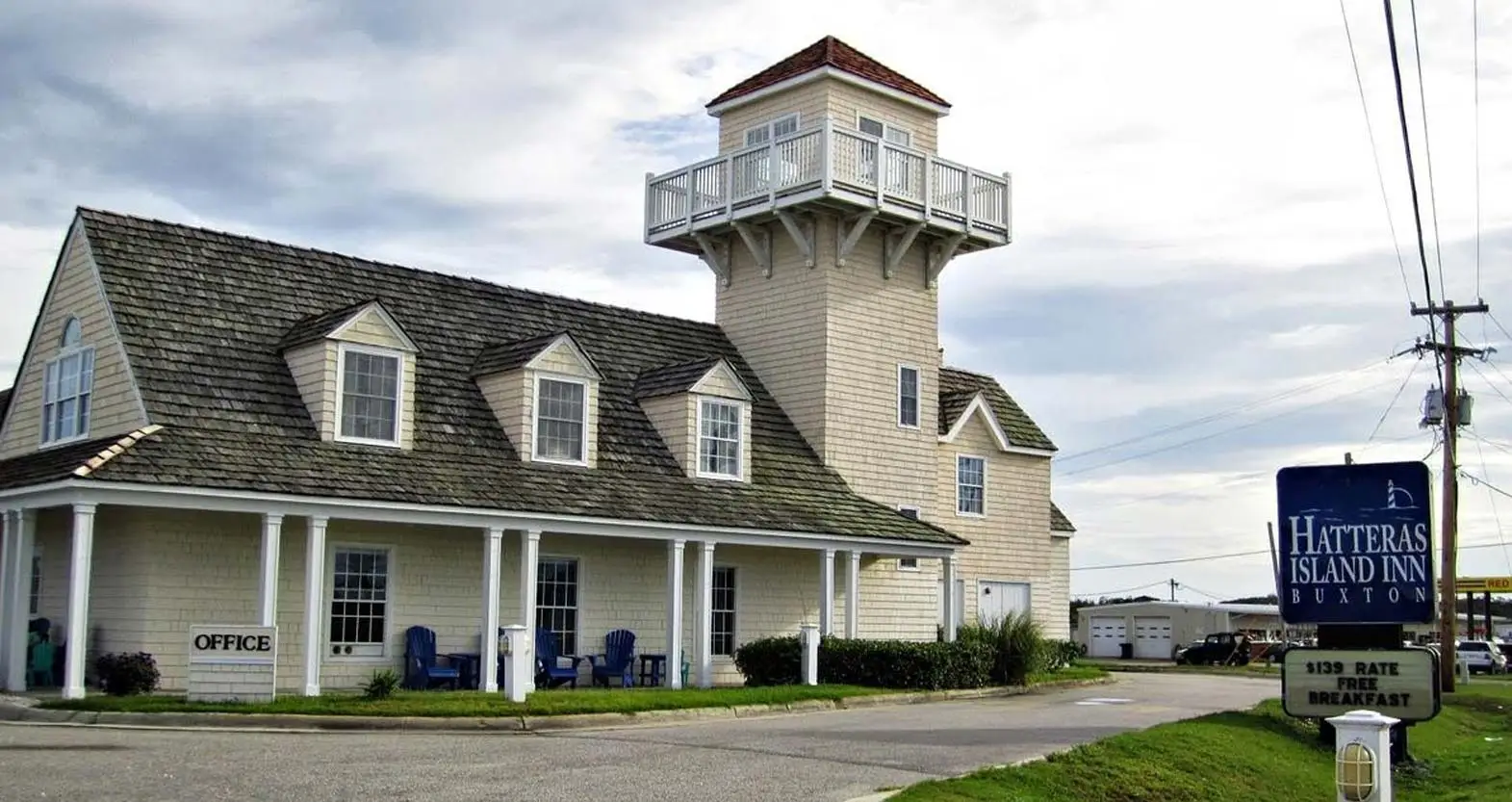 The inn’s shingled tower and porch by daylight under a cloudy sky, with the roadside sign reading “Hatteras Island Inn, Buxton”