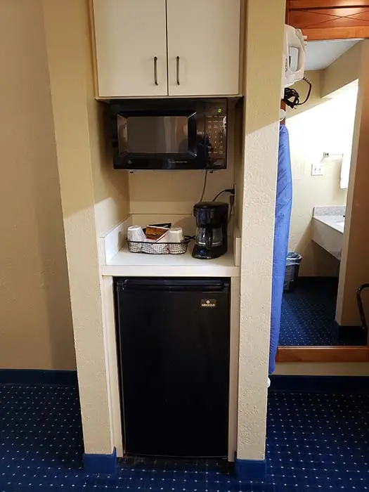 An in-room alcove with a microwave and coffee maker on a shelf above a small black refrigerator